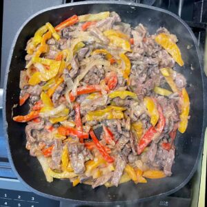 Top down view of a skillet cooking sliced steak, peppers and onions in a cheesy sauce.